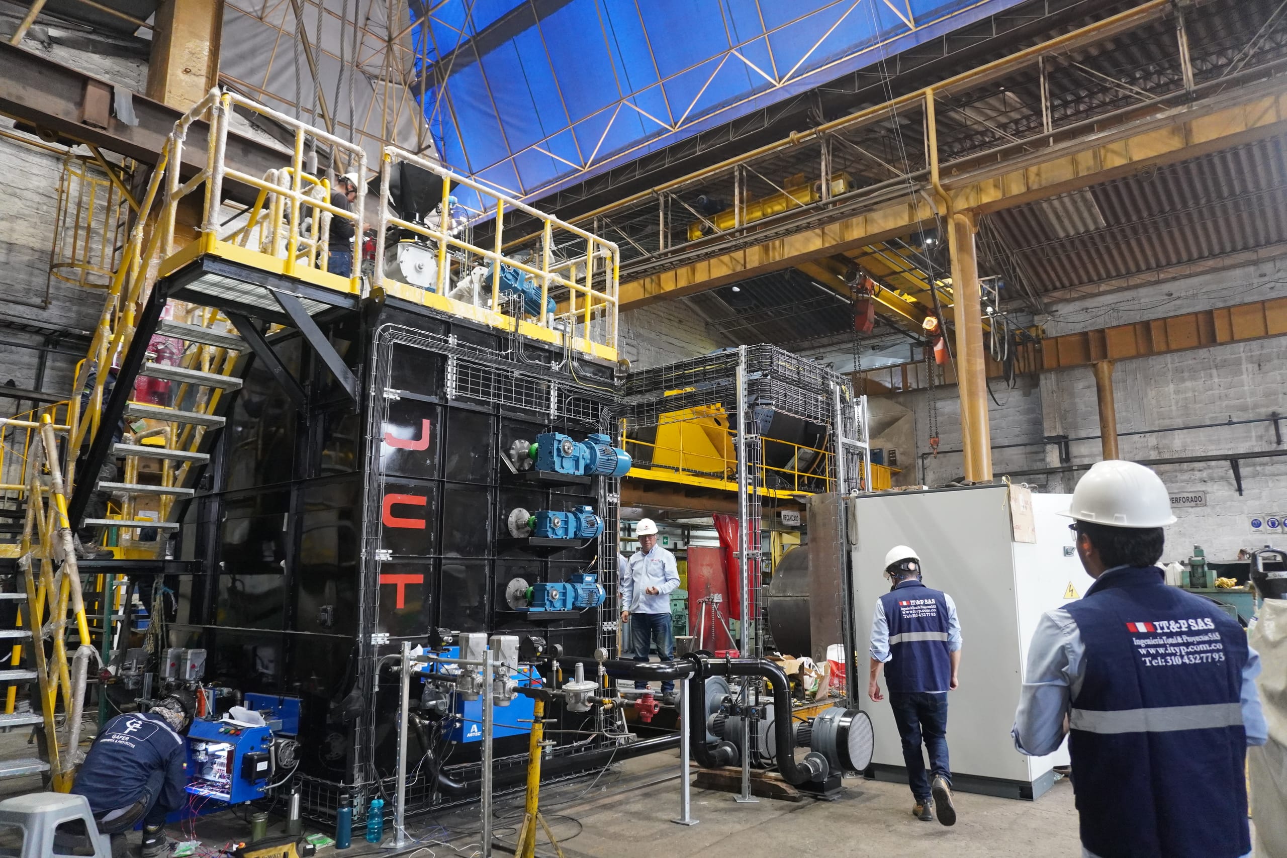 Nave industrial de gran altura con estructura metálica y equipos de proceso; al fondo, dos personas con casco blanco y chaqueta de ITYP.