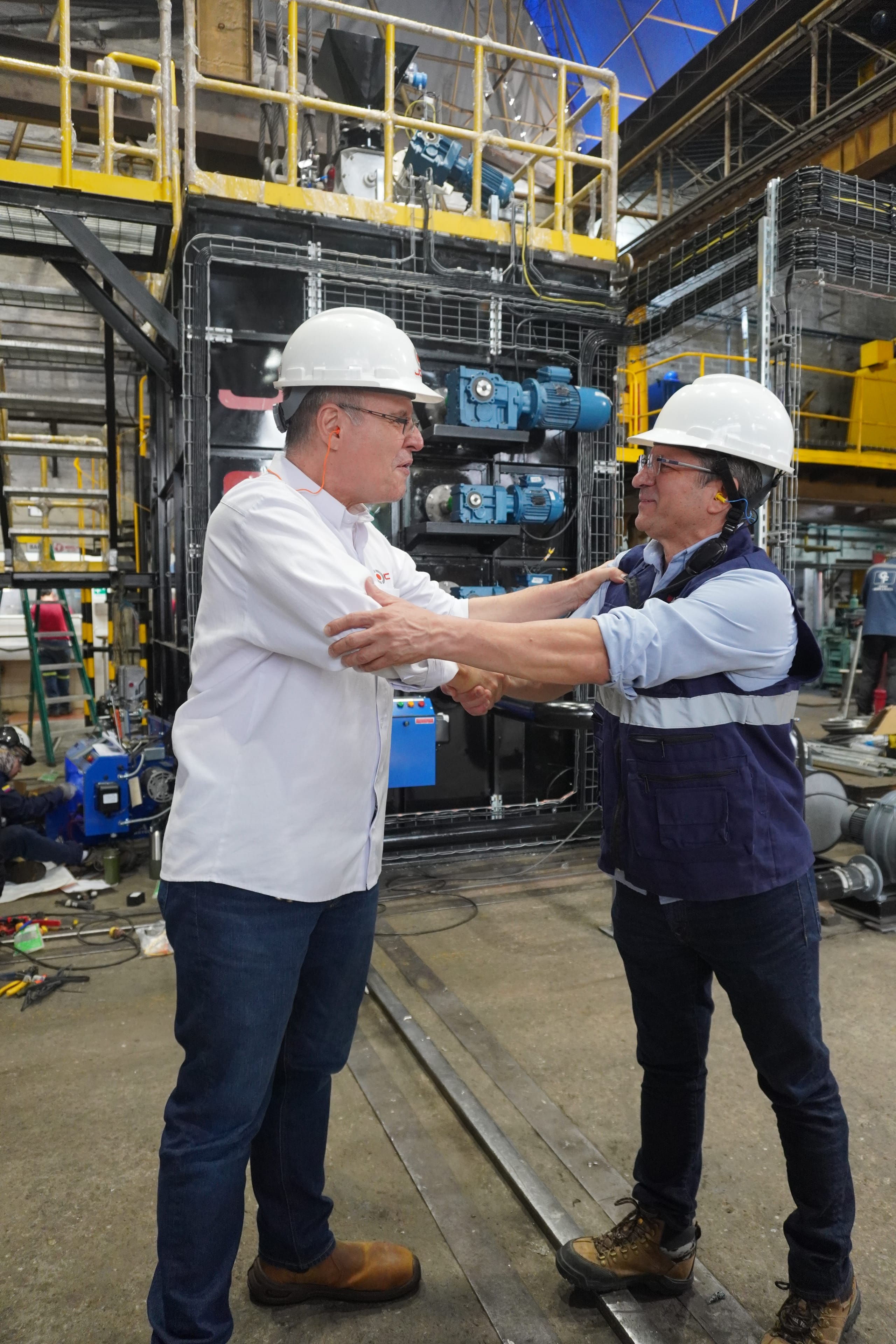 Dos personas con casco blanco se saludan dentro de una planta industrial.