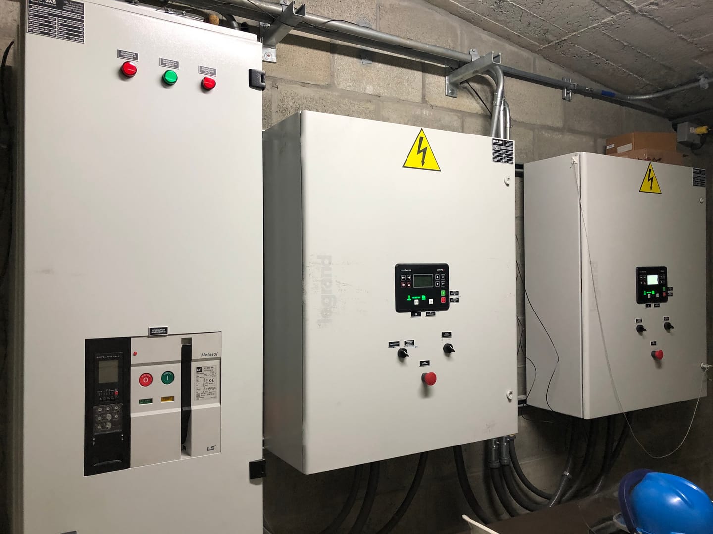 Tres tableros en la subestación: a la izquierda el interruptor de potencia del generador con relé digital de disparo, y a su lado dos tableros de control de sincronismo con controlador de planta eléctrica.
