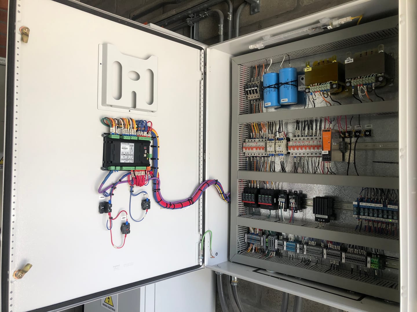 Tablero de control de sincronismo abierto: el controlador de planta eléctrica montado en la puerta y, dentro, protecciones, fuente conmutada, borneras y cableado numerado uno a uno.