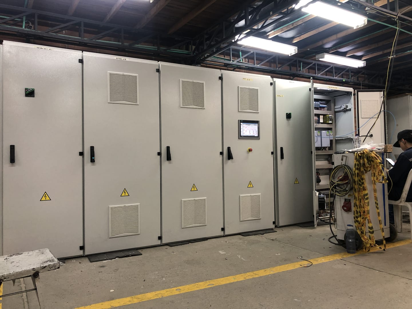 Fila de centros de control de motores instalados en una nave industrial, con personal técnico trabajando al fondo.